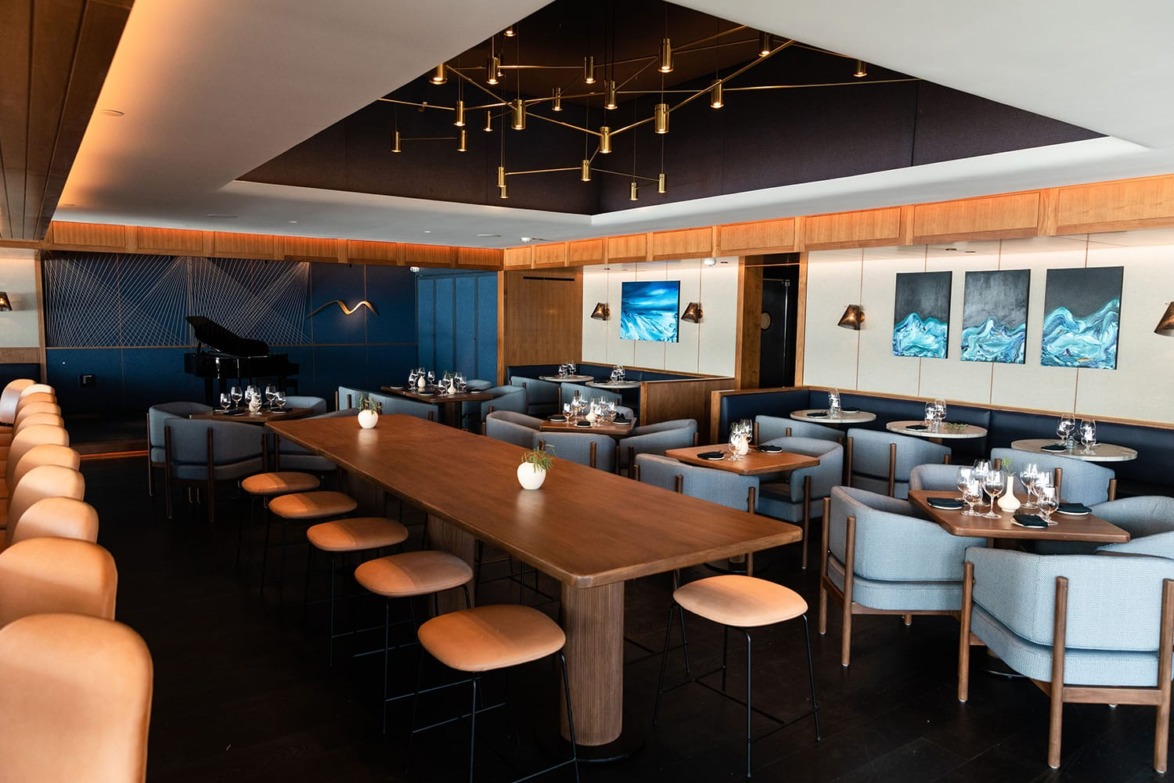 Modern restaurant interior with wooden tables, stylish lighting, blue seating, art on walls, and a grand piano in the corner.