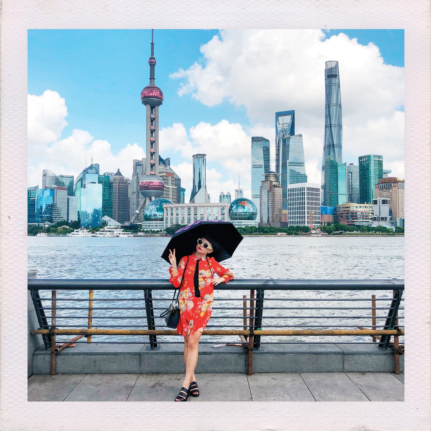 Person posing with umbrella in front of a city skyline featuring tall modern buildings and a river.