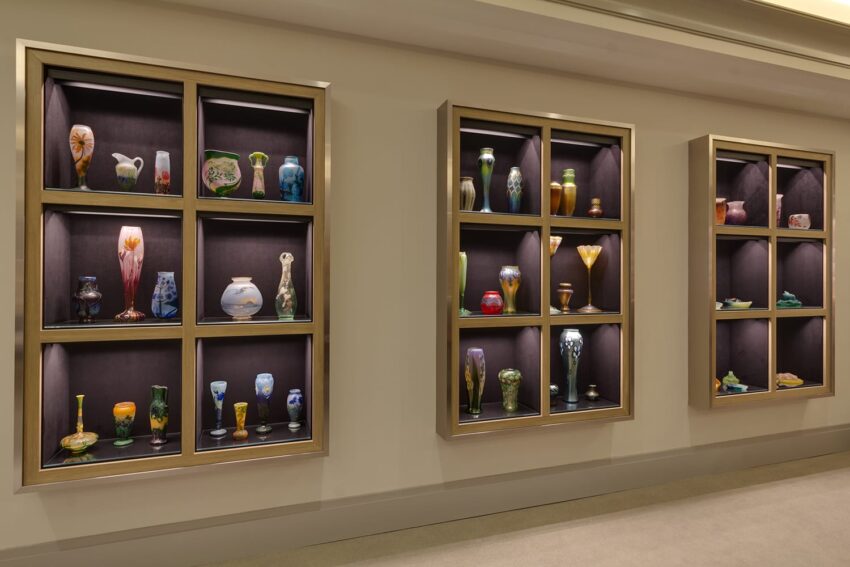 Display of colorful glass vases and sculptures in illuminated wall-mounted shelves at an exhibition.