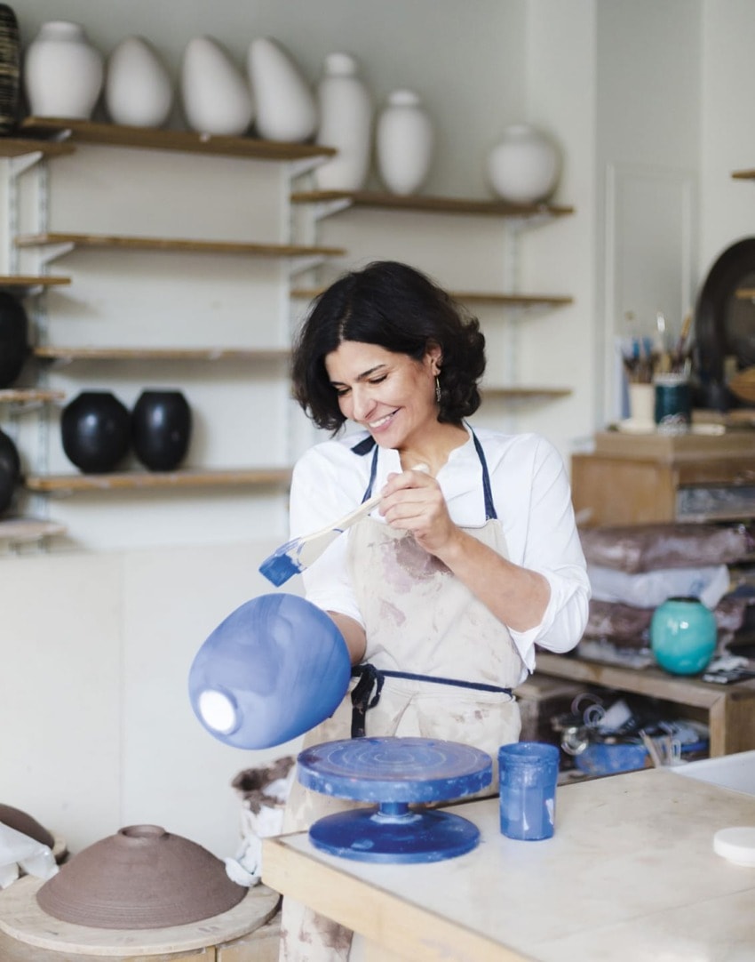 Potter in a workshop painting a blue ceramic vase, surrounded by various pottery pieces on shelves and a table.