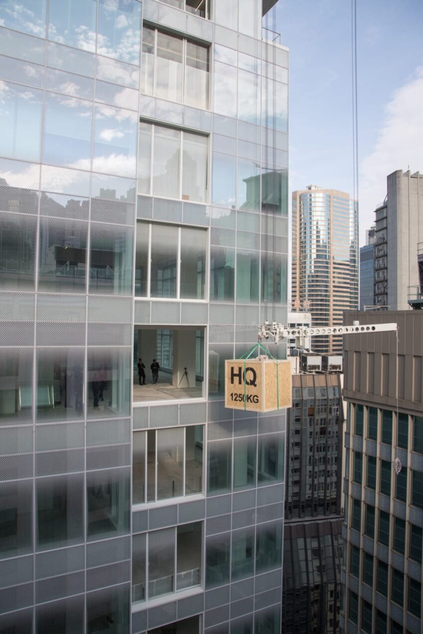 Crate labeled HQ 125KG being lifted by a crane to a high-rise building against a backdrop of cityscape and blue sky.