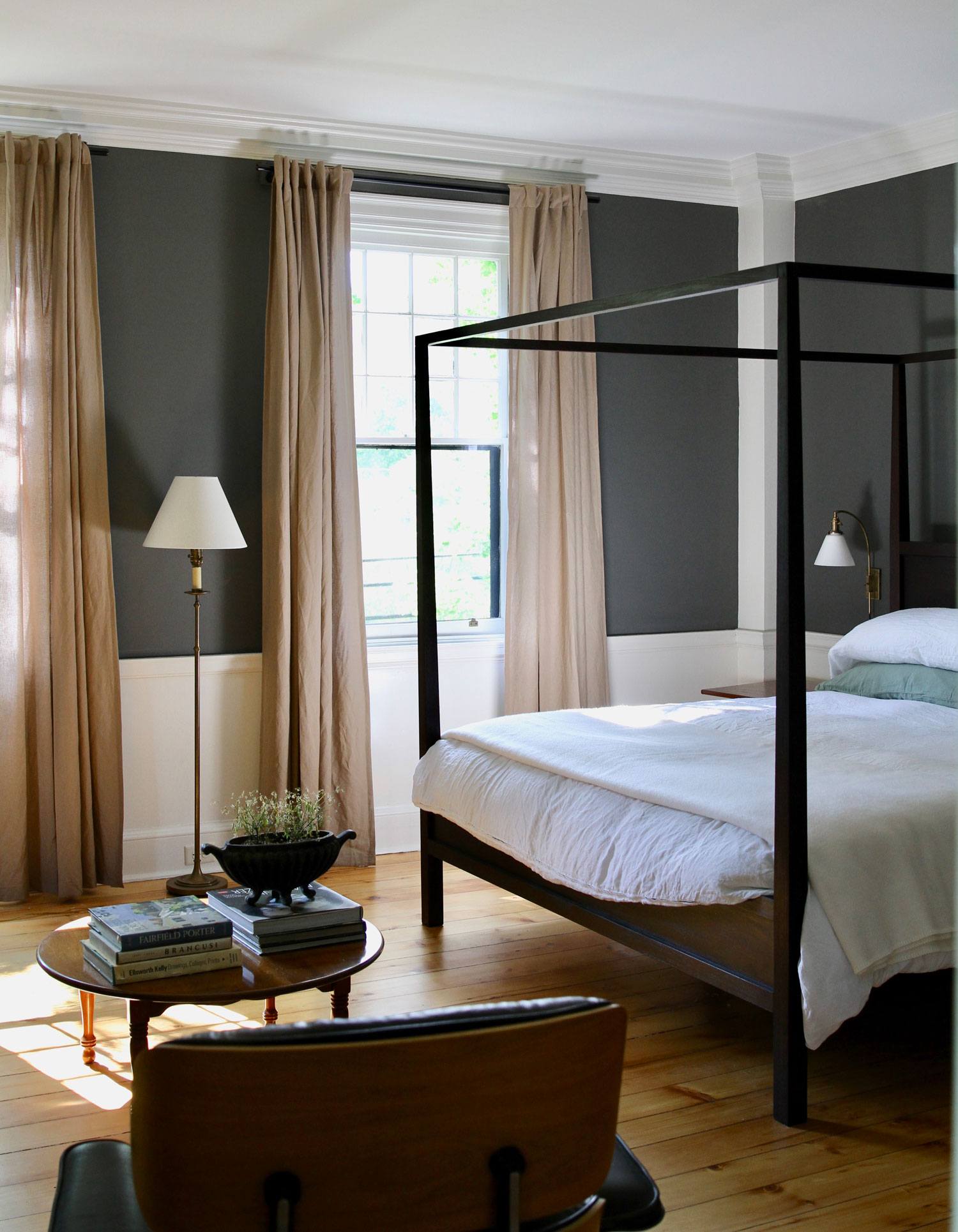 Modern bedroom with four-poster bed, dark walls, large windows, beige curtains, wooden floor, and a lamp on a side table.