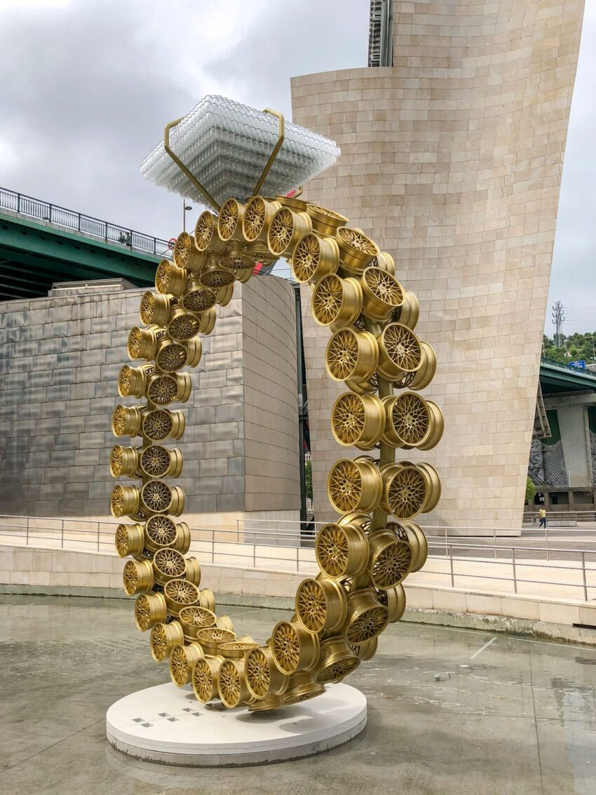 Large sculpture of a diamond ring made from golden car tires with a towering building and bridges in the background.