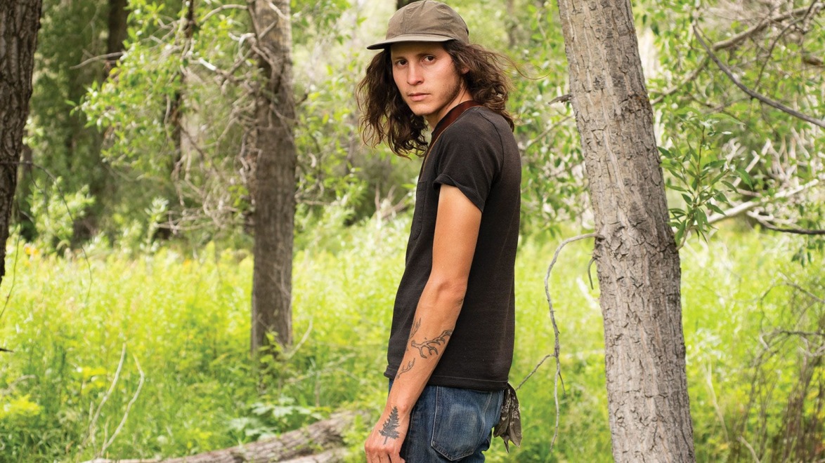 Person with long hair and tattoos standing in a forest, wearing a cap and black t-shirt, looking to the side.