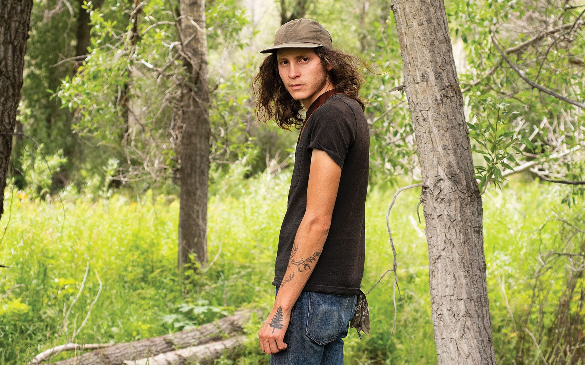 Person with long hair and tattoos standing in a forest, wearing a cap and black t-shirt, looking to the side.