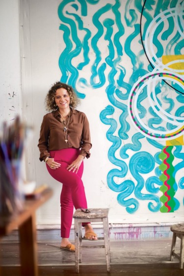 Person standing by a colorful mural with abstract patterns, wearing a brown shirt and pink pants, smiling at the camera.