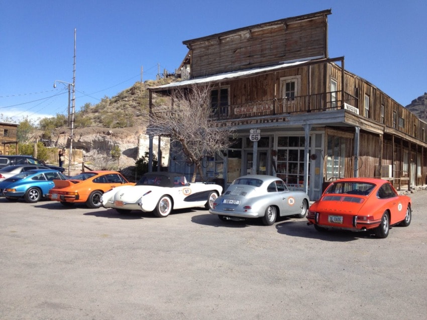Vintage cars parked in front of an old wooden building on a sunny day in a desert town setting.