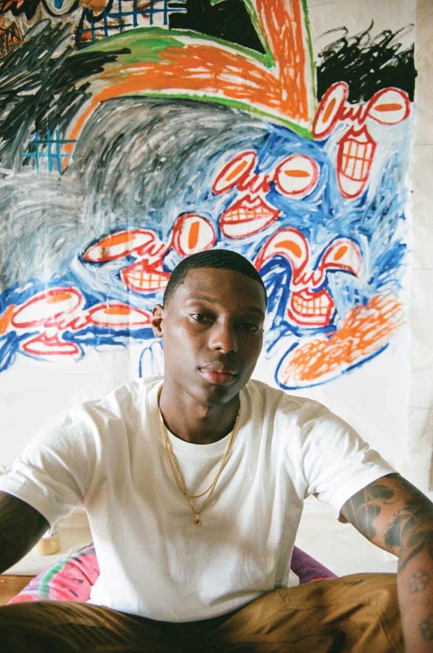 Person in a white shirt sitting in front of a colorful abstract painting with various shapes and patterns.