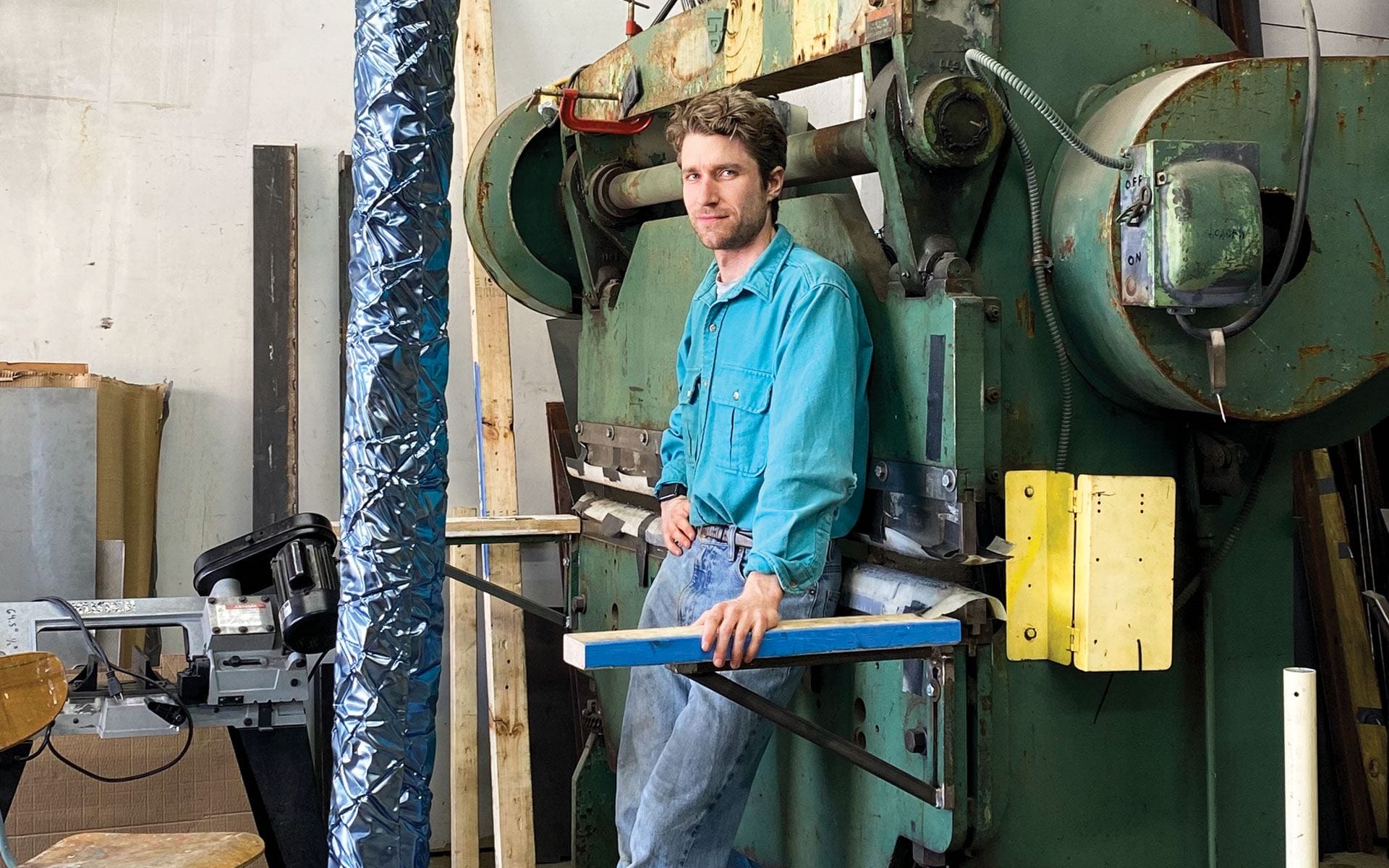 Man in a blue shirt standing by industrial machinery in a workshop environment, holding a tool.