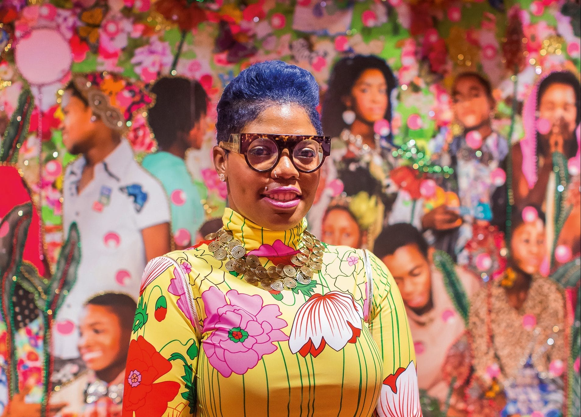Person wearing colorful attire and glasses stands in front of a vibrant, floral background with various portraits.
