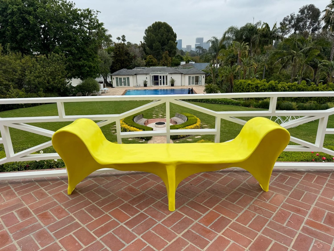 Bright yellow modern bench on terrace overlooking lush garden with pool and trees in the background.