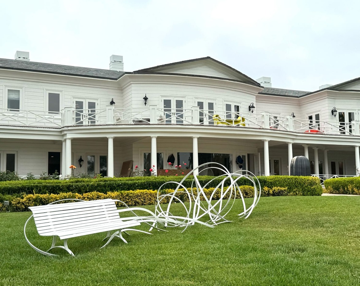 Large white house with unique white metal sculptures on the lawn, featuring an arched balcony and neatly trimmed hedges.