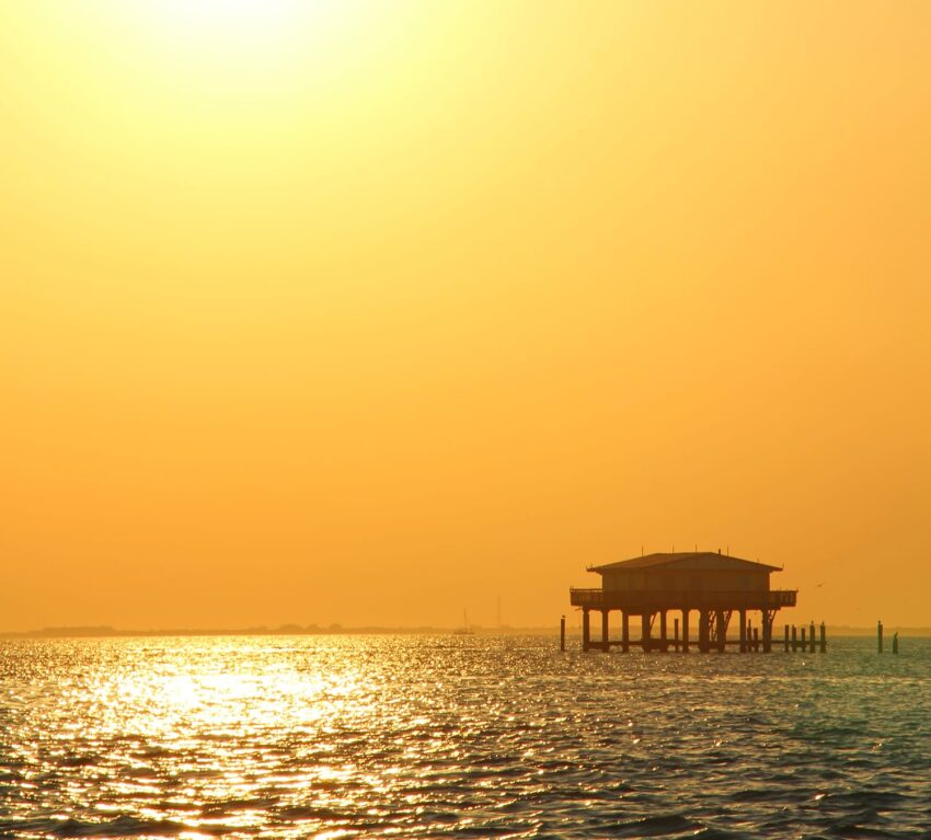 Silhouetted house on stilts over water during a vibrant yellow sunset with calm sea and distant horizon.
