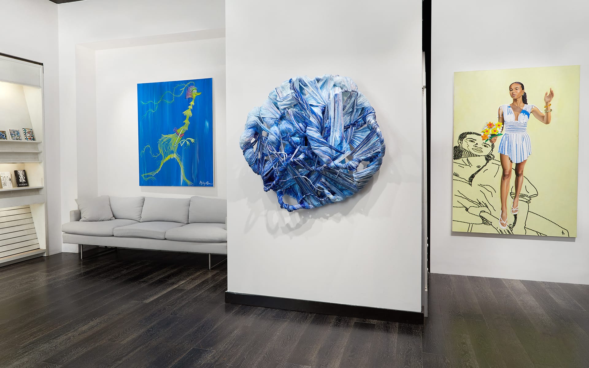 Art gallery interior featuring a sofa, a blue abstract sculpture, and colorful paintings on white walls.