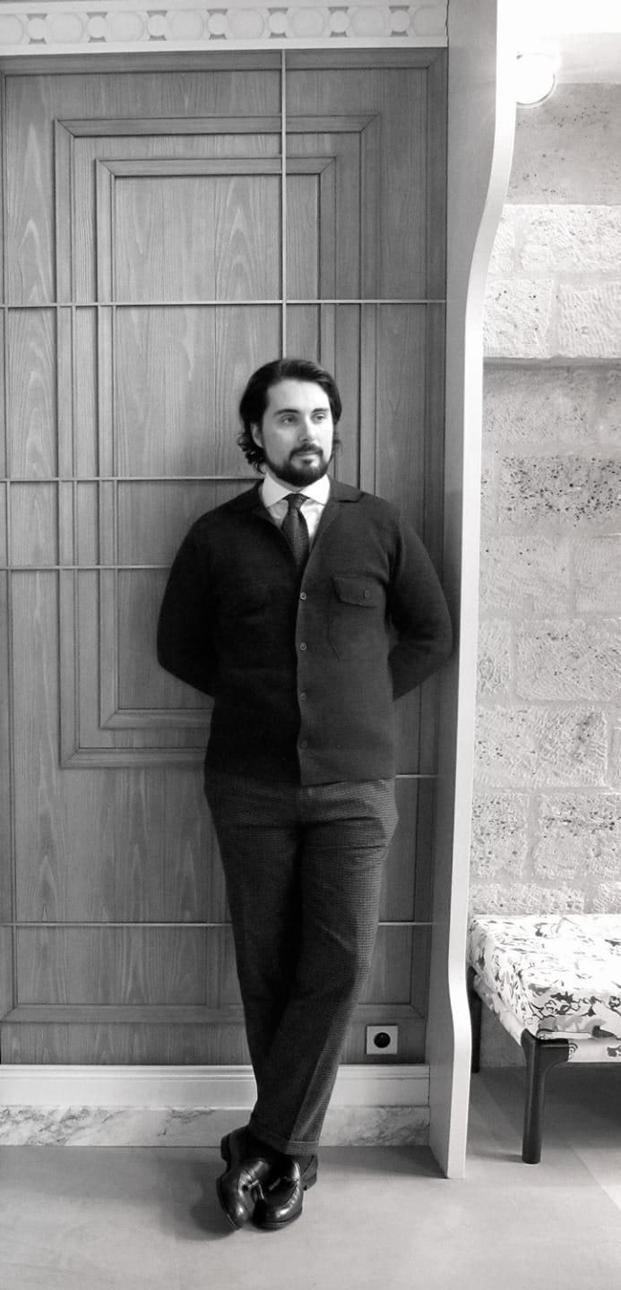 Man standing in front of a wooden door, wearing a suit and tie, with arms behind his back, in a black and white photo.