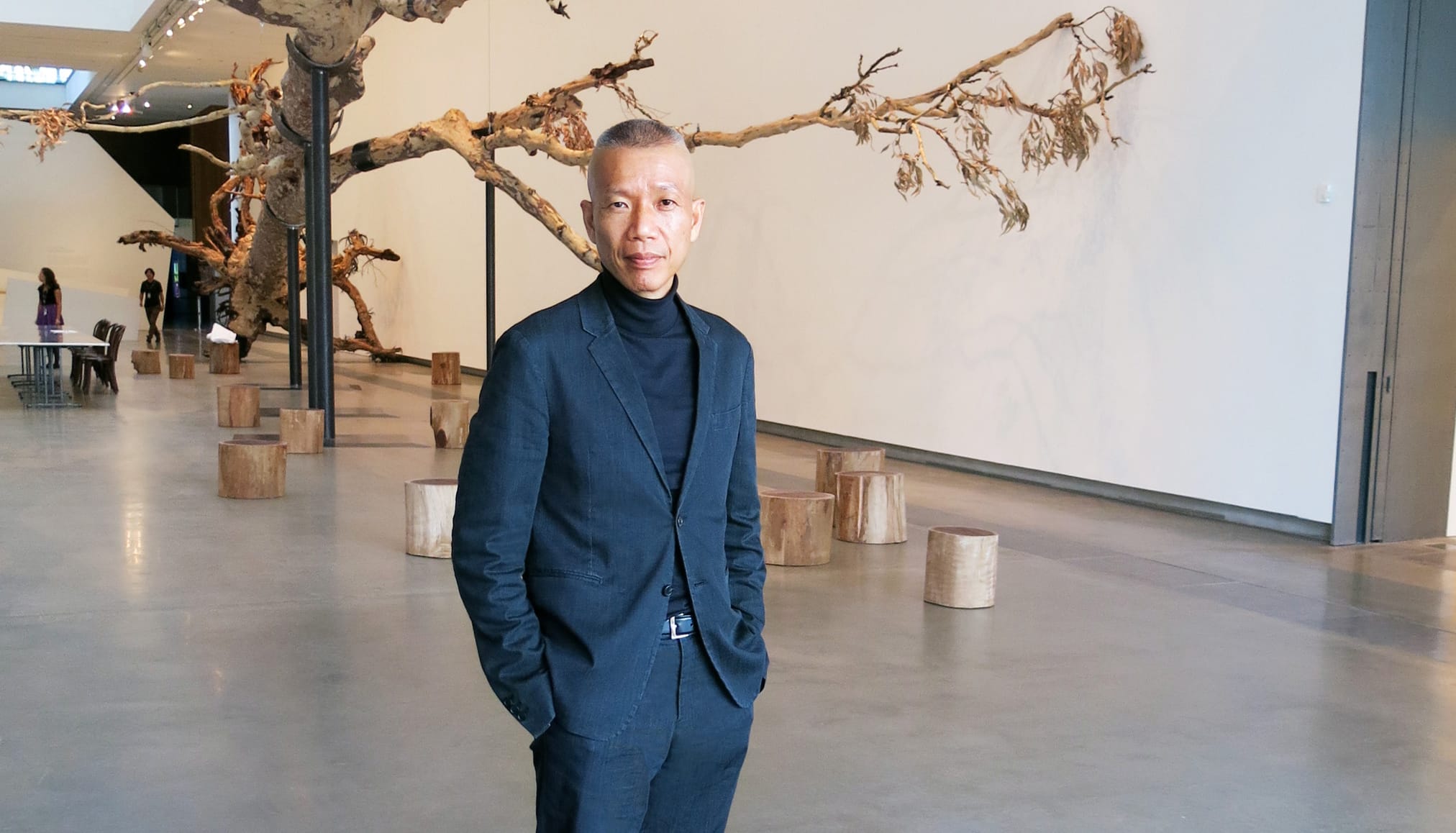 A person in a dark suit stands in a modern art gallery with a large tree sculpture on display in the background.