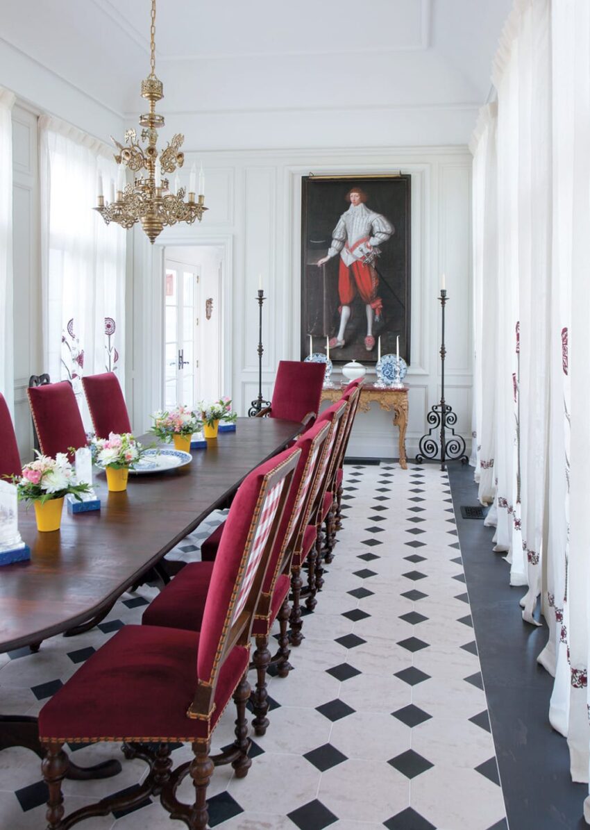 Elegant dining room with red velvet chairs, long wooden table, ornate chandelier, and portrait painting on the wall.