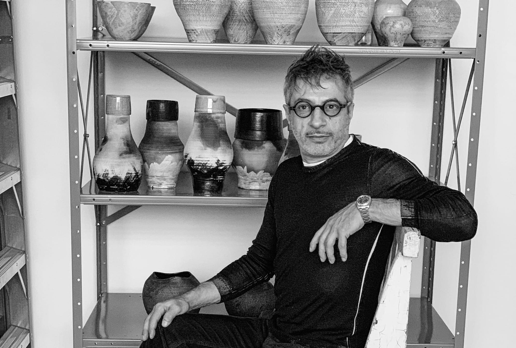 Man sitting in front of shelves with ceramic pots and vases, wearing glasses and a watch, in a monochrome photo.