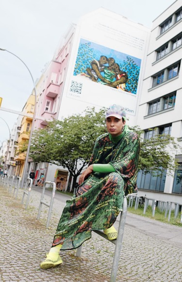 Person in vibrant outfit sitting on a metal fence in an urban area with colorful buildings and wall art in the background