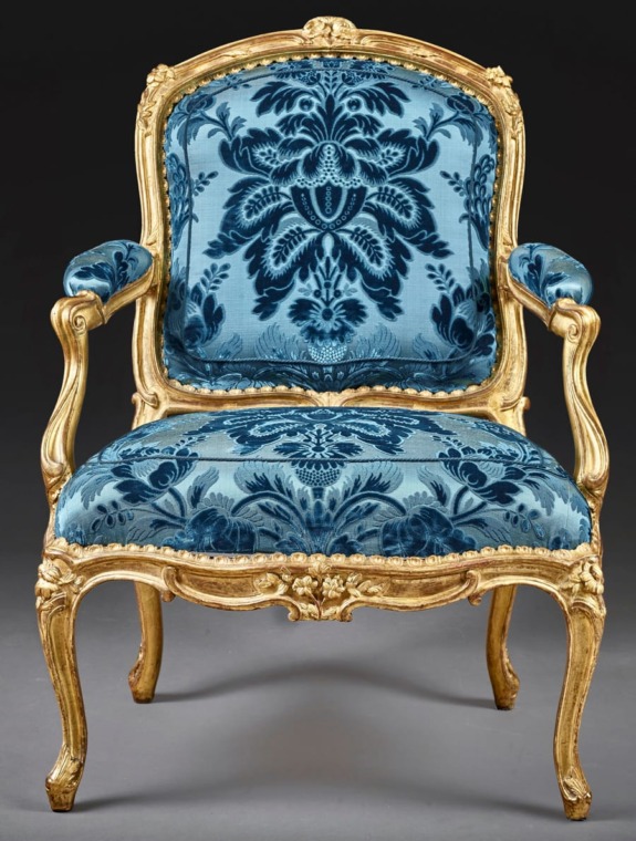 Ornate vintage armchair with gold frame and blue floral upholstery against a dark background.