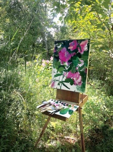 Plein air painting setup in a garden with a colorful floral canvas on an easel surrounded by lush green foliage