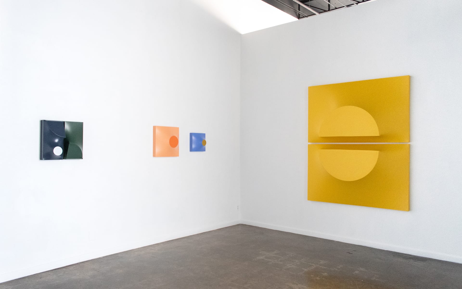 Gallery with abstract geometric art on white walls, featuring green, orange, blue squares, and large yellow circle artwork.