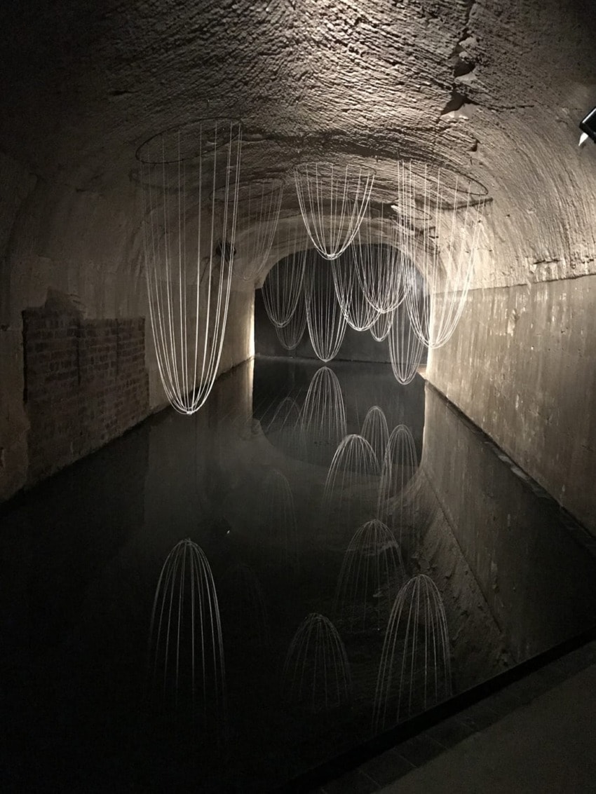 Art installation in a dimly lit tunnel with delicate wire sculptures reflecting on the still water surface below.
