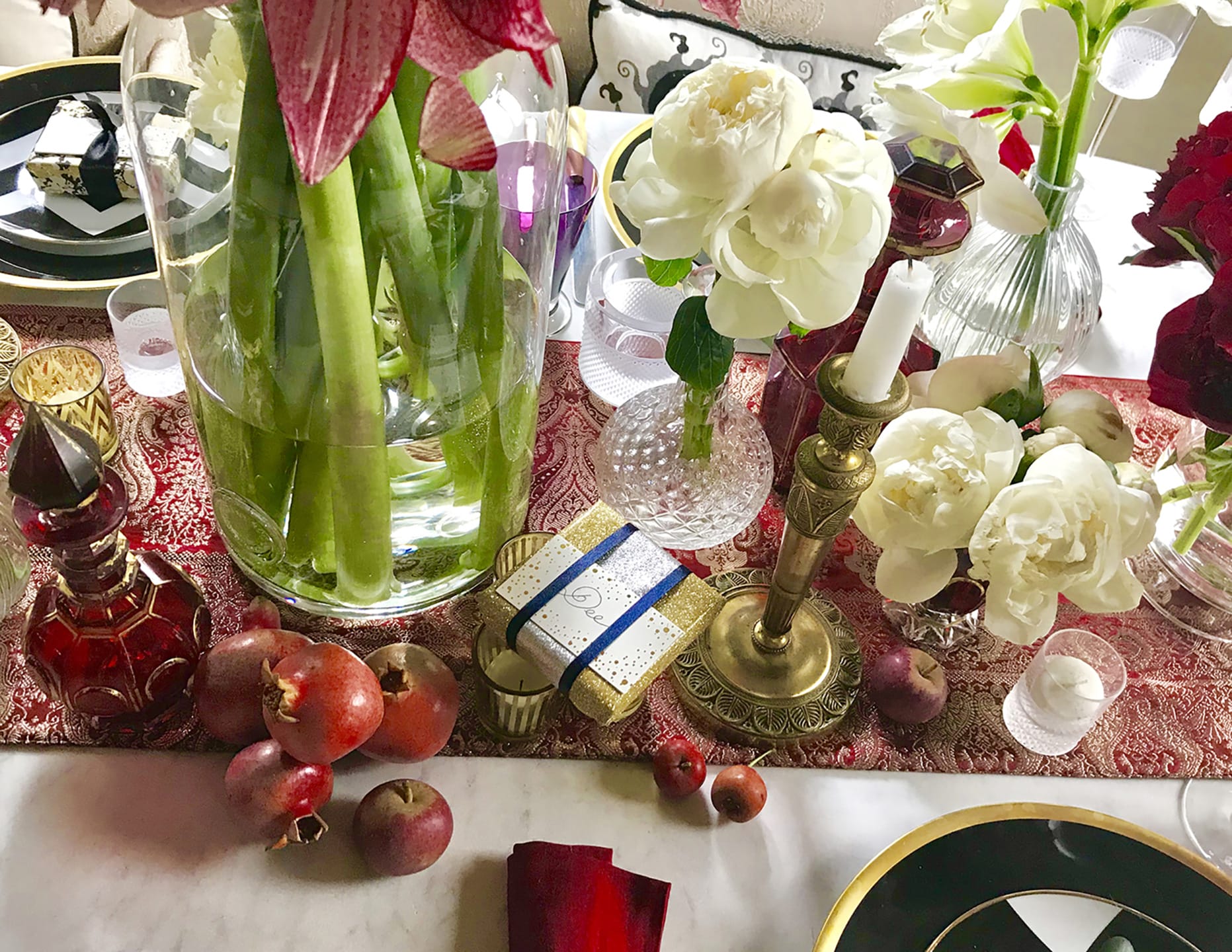 Elegant table setting with flowers, candles, pomegranates, and a wrapped gift on a red tablecloth.