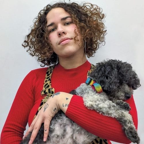 Person with curly hair in red sweater holding a small curly-haired dog wearing a colorful collar against a plain background.
