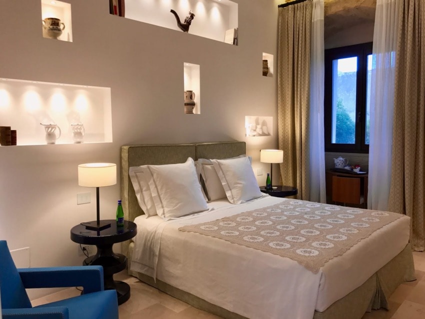 Elegant hotel room with a large bed, bedside tables, decorative shelves, soft lighting, and a window with curtains.