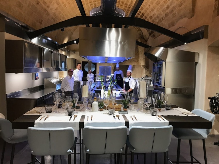 Chefs and staff in a modern kitchen with set dining table, arched ceiling, and stainless steel appliances.