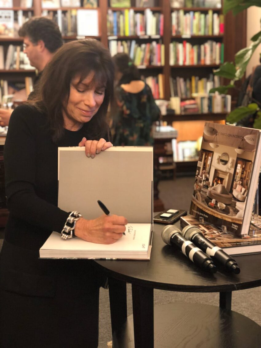 Woman signing a book at a bookstore event, with microphones on a table and bookshelves in the background.