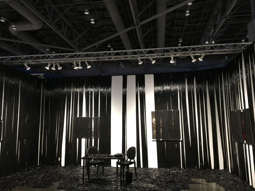 Art installation featuring a room with black and white striped walls, black chairs, and a table, under bright ceiling lights.