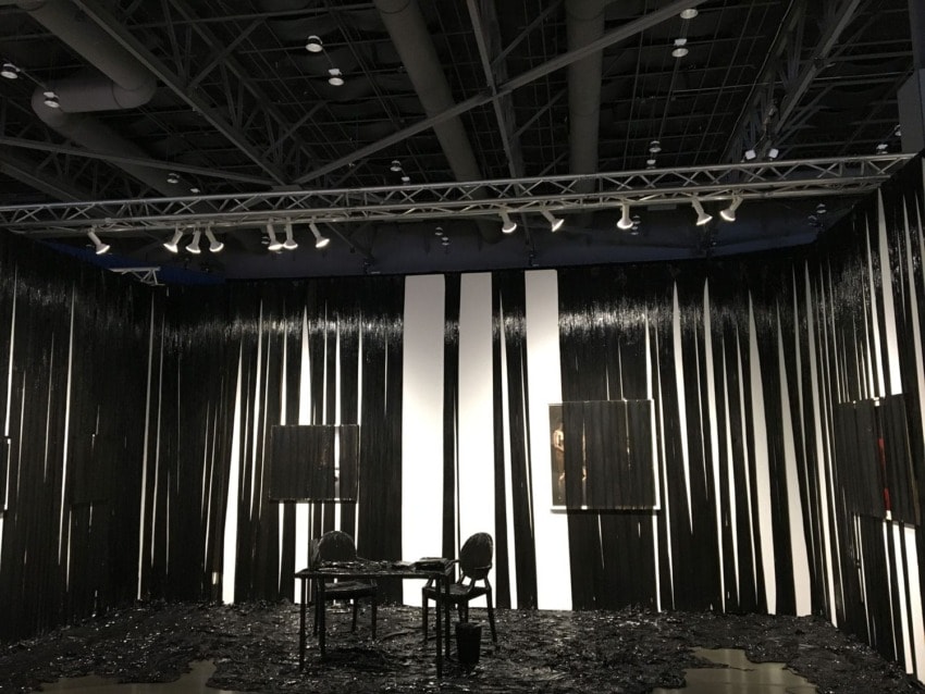 Art installation featuring a room with black and white striped walls, black chairs, and a table, under bright ceiling lights.