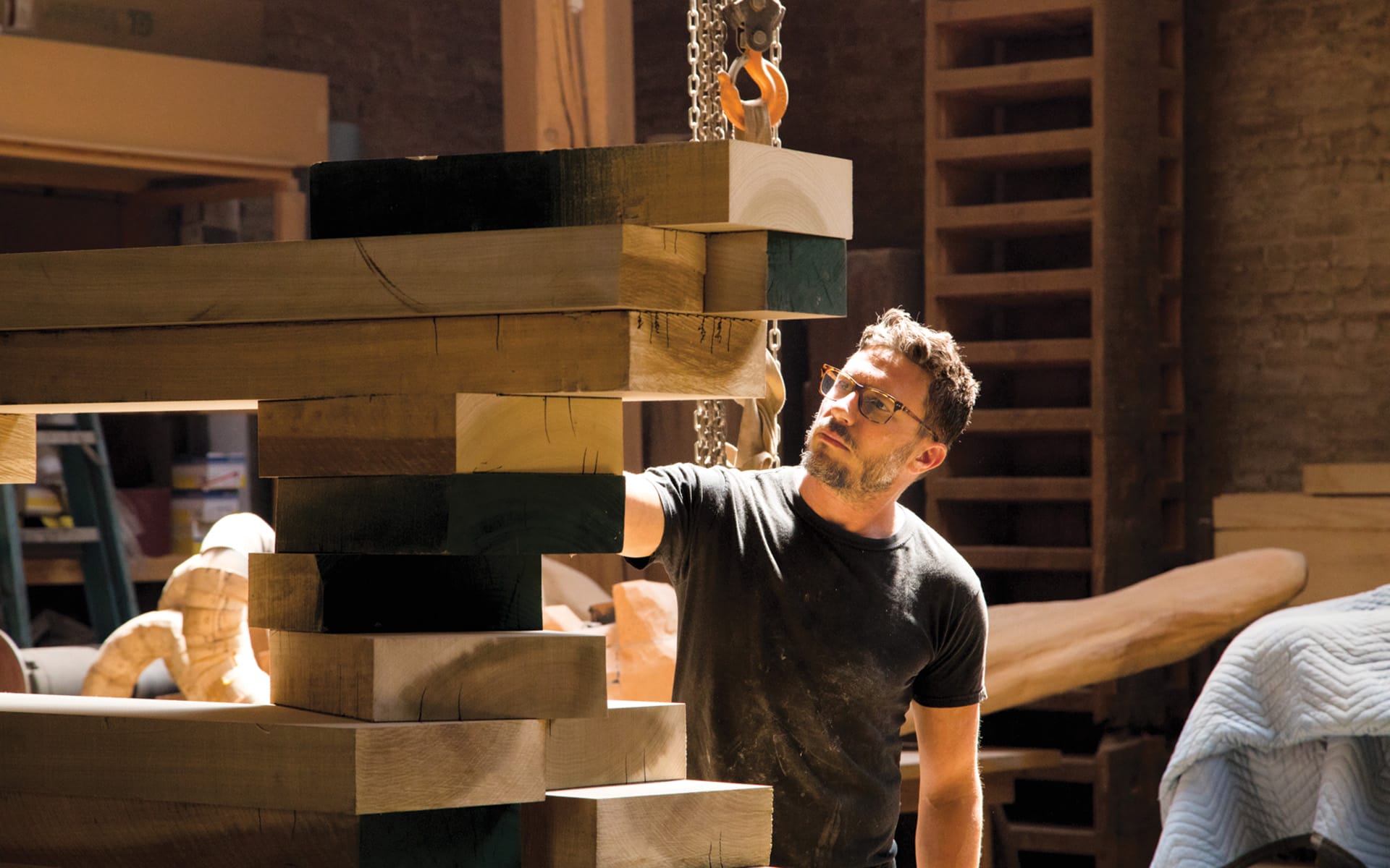 Man adjusting large wooden beams with a pulley system in a workshop filled with natural light.