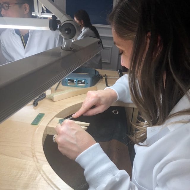 Person in a workshop using tools to craft an object at a workbench under a lamp.