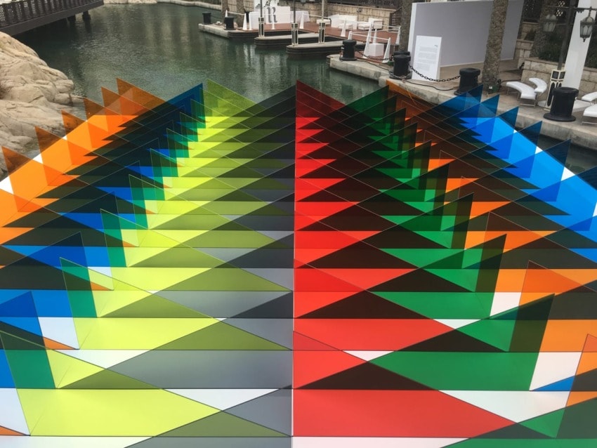 Colorful triangular prism art installation near water with vibrant hues reflecting sunlight.