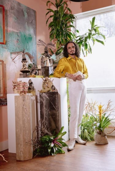 Person in a yellow shirt stands among plants and decorative objects in a sunlit room.