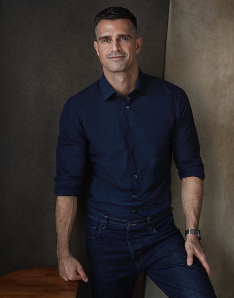 Man in navy shirt and dark jeans smiles while leaning against a wall with one hand on a wooden table.