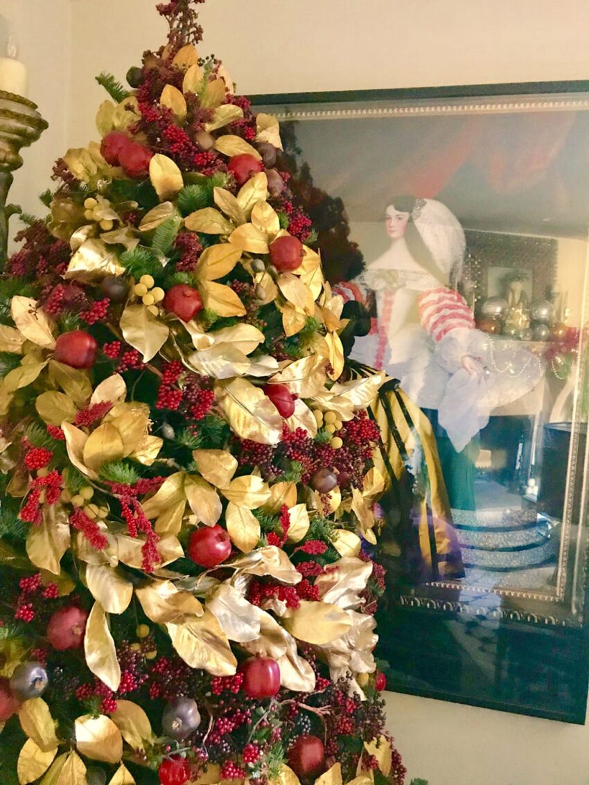 Christmas tree with gold leaves and red berries next to a framed portrait of a historical figure in a decorated room.