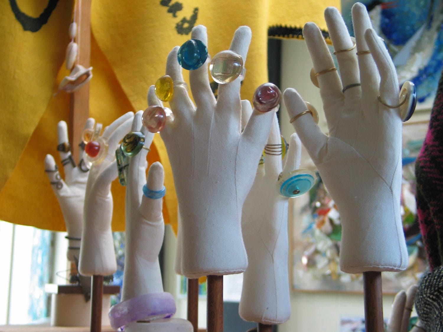 Display of colorful rings on mannequin hands with a yellow fabric backdrop in a jewelry store setting.