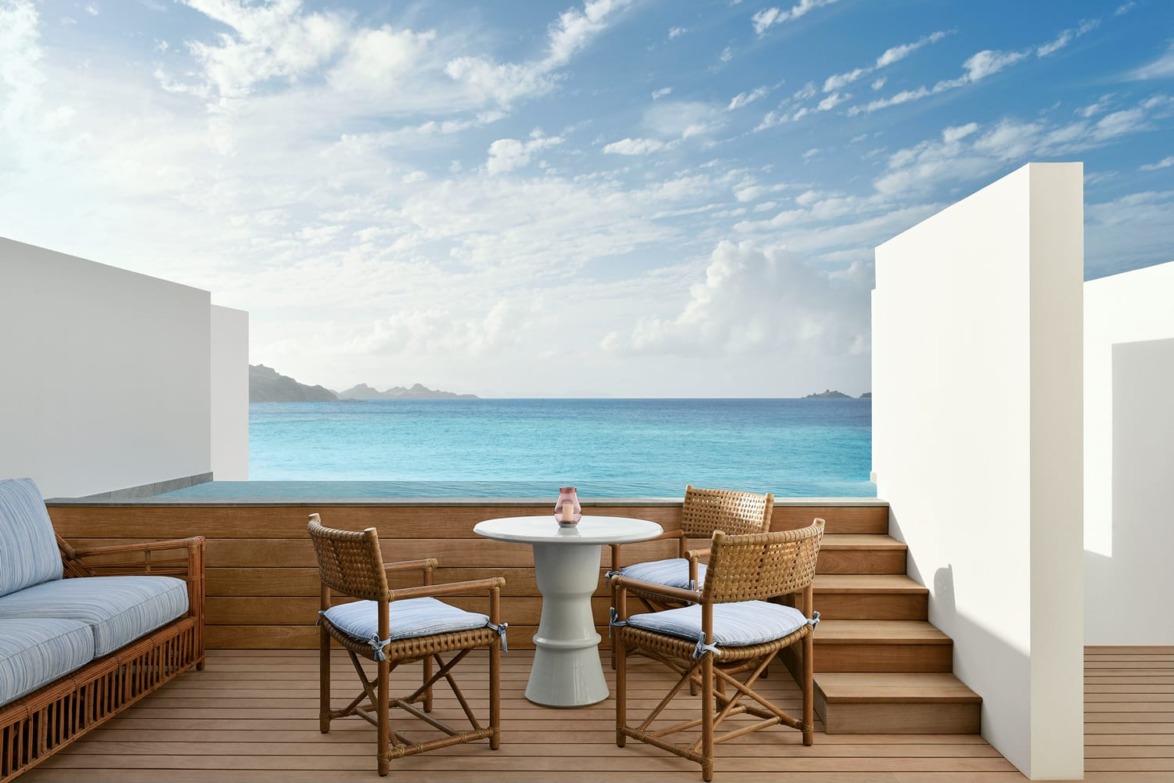 Ocean view from a terrace with wicker chairs and table, wooden decking, and a clear blue sky with scattered clouds.