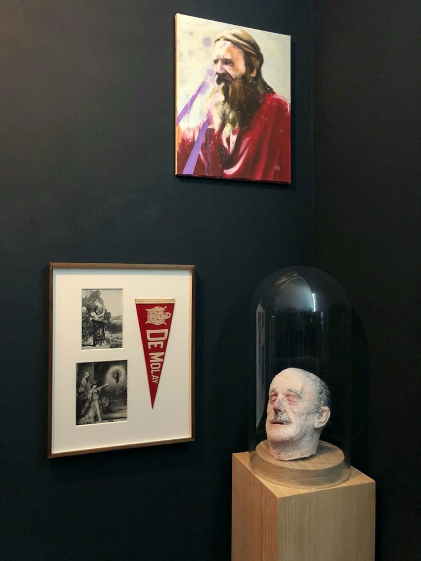 Art gallery corner with a portrait, framed images, red banner, and sculpted head under a glass dome on a wooden stand.