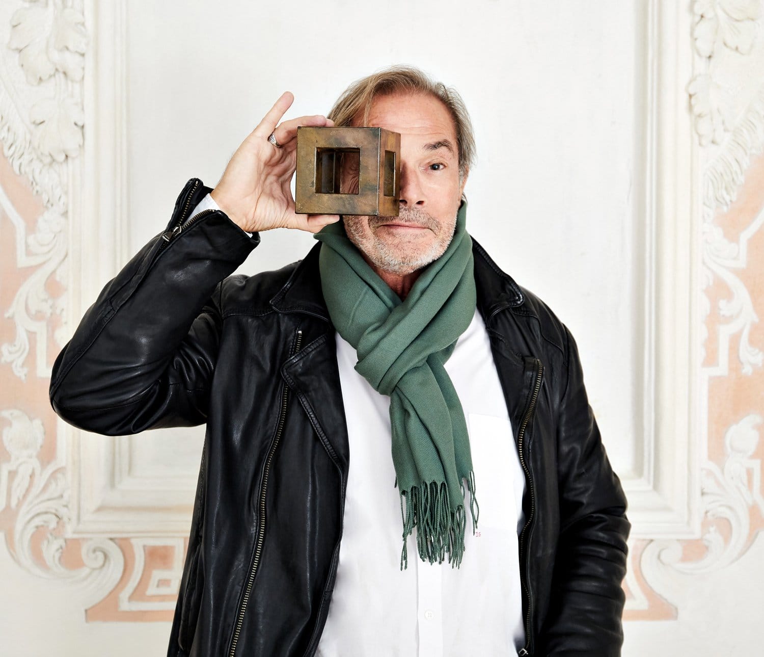 Man in leather jacket and green scarf holding a square object up to his eye against an ornate white background.
