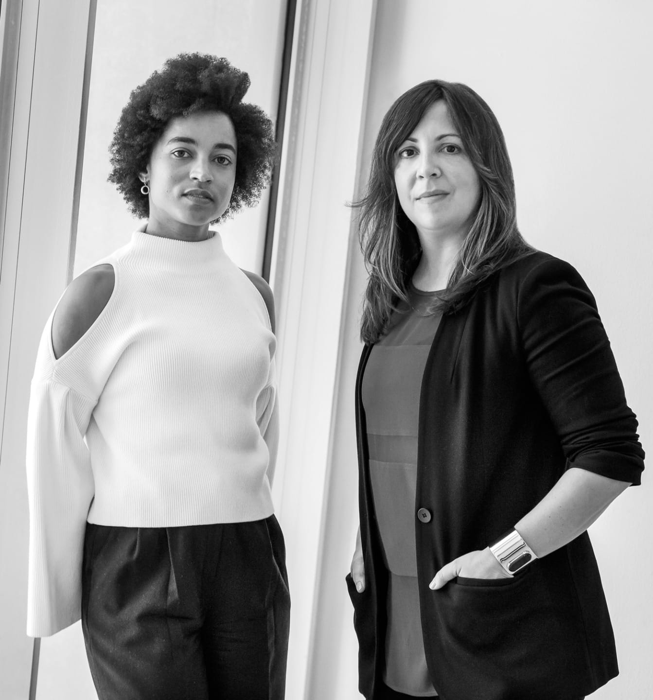 Two people standing by a window, one wearing a white top and black pants, the other in a dark blazer and shirt, in black and white.