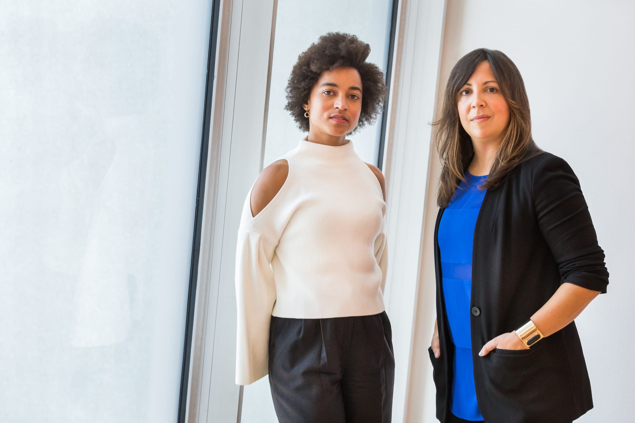 Two women standing by a window, one in a white sweater with open shoulders, and the other in a blue top and black blazer.