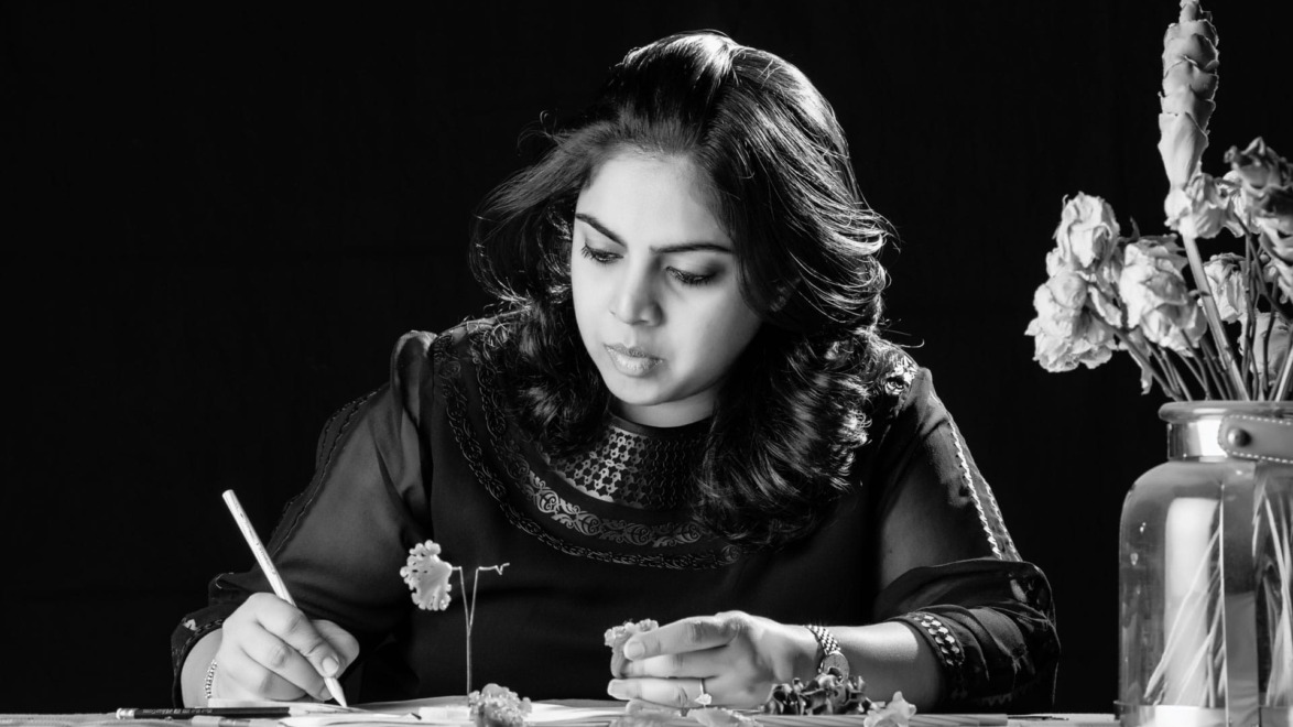 Woman drawing flowers at a table, focused, with a vase of flowers beside her in a black and white setting.