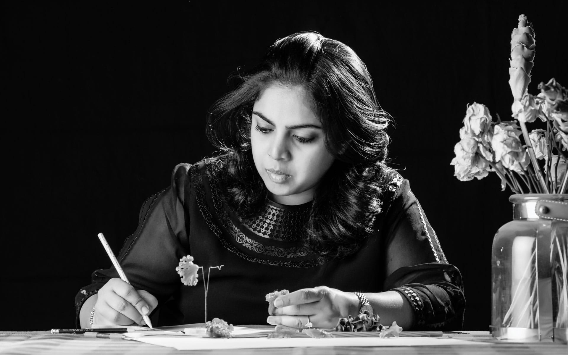 Woman drawing flowers at a table, focused, with a vase of flowers beside her in a black and white setting.