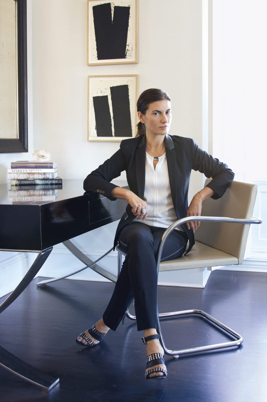 Woman seated at modern desk in stylish office with contemporary artwork on walls, wearing a black blazer and sandals.
