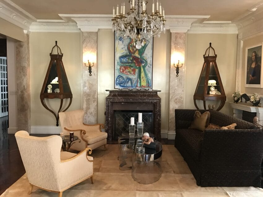 Elegant living room with chandelier, colorful artwork above fireplace, vintage furniture, and wall sconces on marble columns.